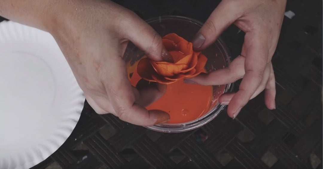 A person dipping a handmade sola wood flower into a bowl of bright orange dye to color the petals